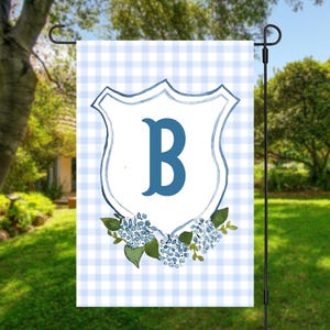 May include: A garden flag with a blue and white gingham pattern. It features a blue shield with the letter "B", surrounded by blue hydrangea flowers and green leaves. The flag is displayed on a black metal stand, suitable for outdoor use.