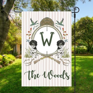 May include: A garden flag with a hunting theme. The flag features a circular design with the letter "W" in the center, flanked by two dogs. The words "The Woods" are written below the circle. The background has vertical stripes.