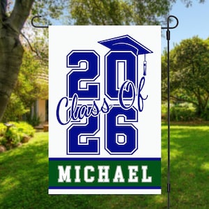 May include: A white garden flag with the text "2026 Class Of" in blue, a graduation cap, and the name "MICHAEL" in white on a green stripe. The flag is displayed outdoors in a garden setting.