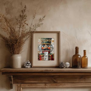 Op de afbeelding: Een ingelijste afbeelding van een kleurrijke, getextureerde voorstelling van een notenkrakersoldaat. De notenkraker heeft een gedetailleerd gezicht met blauwe ogen, een snor en een kroon. Het kunstwerk zit in een houten lijst.