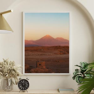 Puede incluir: Una impresión enmarcada de un paisaje montañoso bajo un cielo degradado de tonos azules y naranjas. El primer plano muestra un paisaje desértico con un pequeño edificio. La impresión se exhibe en una estantería de madera con un reloj, un jarrón y una planta.