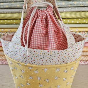 May include: A three-piece fabric basket set. The largest basket is yellow with a white bird pattern. The middle basket has a white background with colorful hearts. The drawstring bag is red and white gingham. The handles are white.