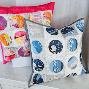 Könnte beinhalten: Zwei dekorative Kissen mit farbenfrohen, kreisförmigen Patchwork-Designs. Ein Kissen hat einen leuchtend rosa Rand und orange, lila und rosa Kreise. Das andere Kissen hat einen grauen Rand und blaue Kreise.