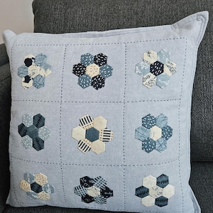 May include: A square, pale blue cushion with a grid pattern. Each square features a flower design made of various blue and white hexagonal fabric pieces. The cushion is on a grey sofa.