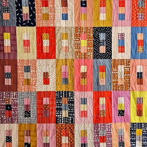 May include: A patchwork quilt with a repeating pattern of rectangular blocks. Each block features a central column of three smaller rectangles in various colours, set against a patterned background. The quilt showcases a variety of colours and textures.