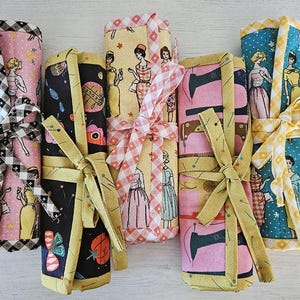 Könnte beinhalten: Sechs gerollte Stoffetuis, jeweils mit einem Band gebunden. Die Muster zeigen Vintage-Frauen, Nähmaschinen und geometrische Designs. Die Farben umfassen Rosa, Gelb, Schwarz und Blau, mit kontrastierenden Schleifen.