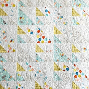May include: A white patchwork quilt with a repeating pattern. The quilt features squares with yellow striped triangles, and sections with colourful polka dots and whimsical patterns in blue, orange, and grey. The quilt has a quilted texture.