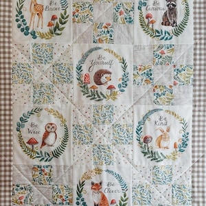 May include: A patchwork quilt with woodland animal illustrations and motivational text. Each square features a different animal, including a deer, raccoon, and fox, within a floral wreath. Phrases such as "Be Brave" and "Be Kind" are displayed.