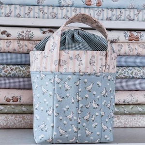 May include: A blue fabric tote bag with a duck and floral pattern. The top edge features a pink and white striped fabric with bunny illustrations. The interior is a blue and white gingham pattern. The bag has a matching handle.