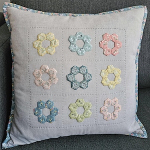 Puede incluir: Cojín decorativo cuadrado con una funda de lino gris claro. Nueve aplicaciones florales coloridas están dispuestas en un patrón de cuadrícula. Las flores son en tonos de amarillo, rosa, azul y verde. El cojín tiene un borde floral.