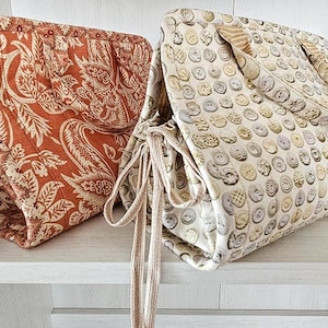 Könnte beinhalten: Zwei Stofftaschen mit Kordelzugverschlüssen. Eine Tasche hat ein rostfarbenes Paisley-Muster, die andere ein Muster aus verschiedenen Knöpfen. Beide Taschen haben passende Stoffgriffe und Kordelzüge.