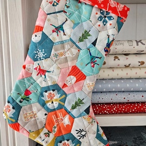 Peut inclure: Une chaussette de Noël matelassée avec un motif patchwork. Le bord est rouge avec un motif de feuilles blanches. Le corps est composé de pièces de tissu hexagonales avec des illustrations sur le thème de l'hiver, dont des skieurs, des arbres et des flocons de neige.