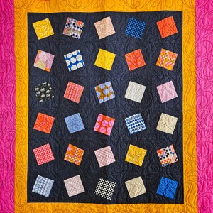 May include: A handmade quilt with a dark blue centre featuring a pattern of colourful square patches. The quilt has a yellow and orange border, and a pink outer border with a swirling pattern. The squares are various colours and patterns.