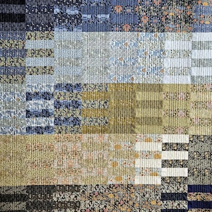 Könnte beinhalten: Ein Patchwork-Quilt mit einer Vielzahl von floralen und geometrischen Mustern in Blau-, Grün-, Gold- und Schwarztönen. Der Quilt besteht aus rechteckigen Blöcken, wobei vertikale und horizontale Linien einen strukturierten Effekt erzeugen. Das Gesamtdesign ist eine Mischung aus traditionellen und modernen Stilen.
