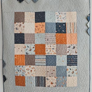May include: A patchwork quilt with a variety of patterned squares in shades of blue, orange, brown, and cream. The quilt has a light blue border and is hanging from a metal rod. The quilt's design includes floral, striped, and text patterns.