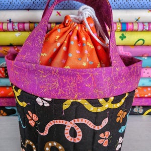 Puede incluir: Un bolso tote hecho a mano con un exterior acolchado negro con coloridas serpientes y mariposas. La parte superior y el asa son de un morado vibrante con detalles dorados. En el interior hay una bolsa con cordón naranja con formas geométricas.