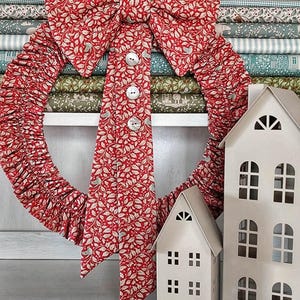 Op de afbeelding: Een rode stoffen krans met een wit en beige bladerpatroon, versierd met een grote strik en witte knopen. Twee witte decoratieve huizen staan voor gestapelde stoffen rollen.