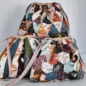 Puede incluir: Tres bolsas con cordón hechas de un mosaico de piezas de tela con varios patrones florales y geométricos. Las bolsas presentan una paleta de colores rojo oscuro, azul marino, verde oliva, crema y marrón, con cordones de cinta claros.