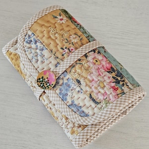 Può includere: Un astuccio in tessuto trapuntato con un design patchwork floreale. L'astuccio presenta una chiusura con bottone e un bordo a quadretti. Il patchwork include motivi floreali nei toni del rosa, blu e giallo. L'astuccio è chiuso e sembra piegato.
