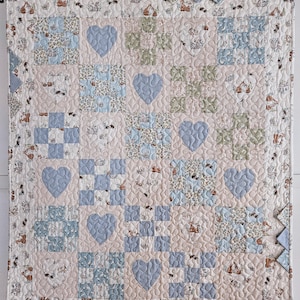 May include: A square patchwork quilt featuring a repeating grid of alternating blocks. The design includes light blue gingham hearts, sage green and floral patterned squares, and a cream-colored background fabric printed with small brown rabbits.