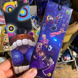 May include: Two illustrated bookmarks. One has a black design with a bear-like face and text. The other is purple with cartoon characters. Each has a hole at the top for a string, ideal for marking pages.