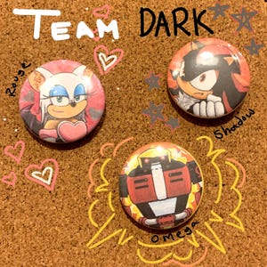 May include: Three circular badges featuring characters from the Sonic the Hedgehog series. The badges depict Rouge, Shadow, and Omega, with the words "Team Dark" above. The background is a corkboard with drawn hearts and stars.