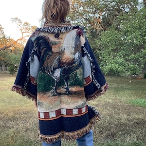 以下が含まれることがあります： 大きな雄鶏のデザインが施された濃い青色のタペストリーコート。コートはフリンジの裾と袖があり、下部に赤と白の正方形のパターンが繰り返されています。雄鶏は茶色、白、黒の色合いで描かれています。
