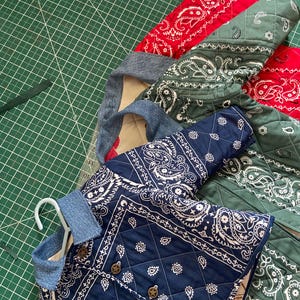 May include: A partially constructed quilted jacket in navy blue, red, and green paisley patterns. The jacket features star-shaped buttons and a denim collar. The pieces are laid out on a green cutting mat.