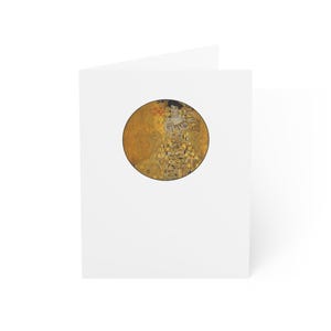 May include: A white greeting card featuring a circular print of Gustav Klimt's "Portrait of Adele Bloch-Bauer I." The artwork depicts a woman in a gold and patterned dress, set against a gold background. The card is folded.