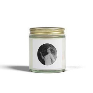 Pode incluir: Um frasco de vela de vidro transparente com uma tampa de metal dourada. A parte frontal do frasco apresenta um rótulo quadrado branco com uma fotografia circular a preto e branco de uma pessoa com uma blusa sem mangas a olhar para cima.