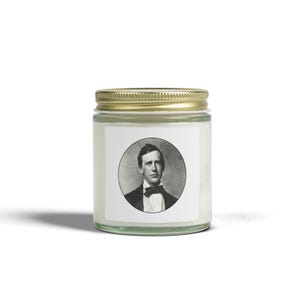 Peut inclure: Un pot à bougie en verre transparent avec un couvercle en métal doré. Le pot présente une étiquette carrée blanche avec un portrait circulaire en noir et blanc d'un homme portant un costume et un nœud papillon centré sur le verre.
