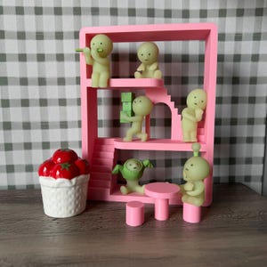 K&ouml;nnte beinhalten: Ein rosa Miniaturhaus mit mehreren kleinen, hellgr&uuml;nen Figuren in verschiedenen Posen. Ein wei&szlig;er Keramikkorb mit roten Tomaten befindet sich links. Ein kleiner rosa Tisch und Hocker befinden sich im Vordergrund.