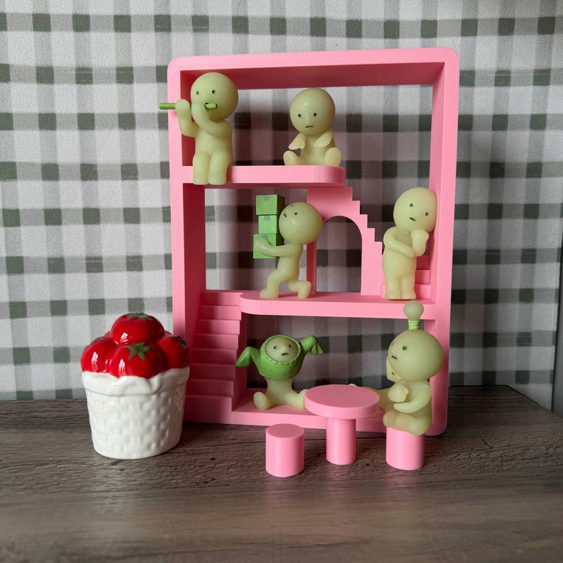 K&ouml;nnte beinhalten: Ein rosa Miniaturhaus mit mehreren kleinen, hellgr&uuml;nen Figuren in verschiedenen Posen. Ein wei&szlig;er Keramikkorb mit roten Tomaten befindet sich links. Ein kleiner rosa Tisch und Hocker befinden sich im Vordergrund.