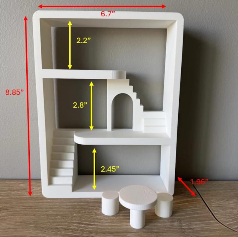 K&ouml;nnte beinhalten: Ein wei&szlig;es Miniatur-Puppenhaus mit rechteckigem Rahmen, das mehrere Ebenen, Treppen und einen gew&ouml;lbten T&uuml;rrahmen aufweist. Enth&auml;lt einen kleinen Tisch und zwei Hocker. Das Puppenhaus ist 22,48 cm hoch, 17,02 cm breit und 4,98 cm tief.