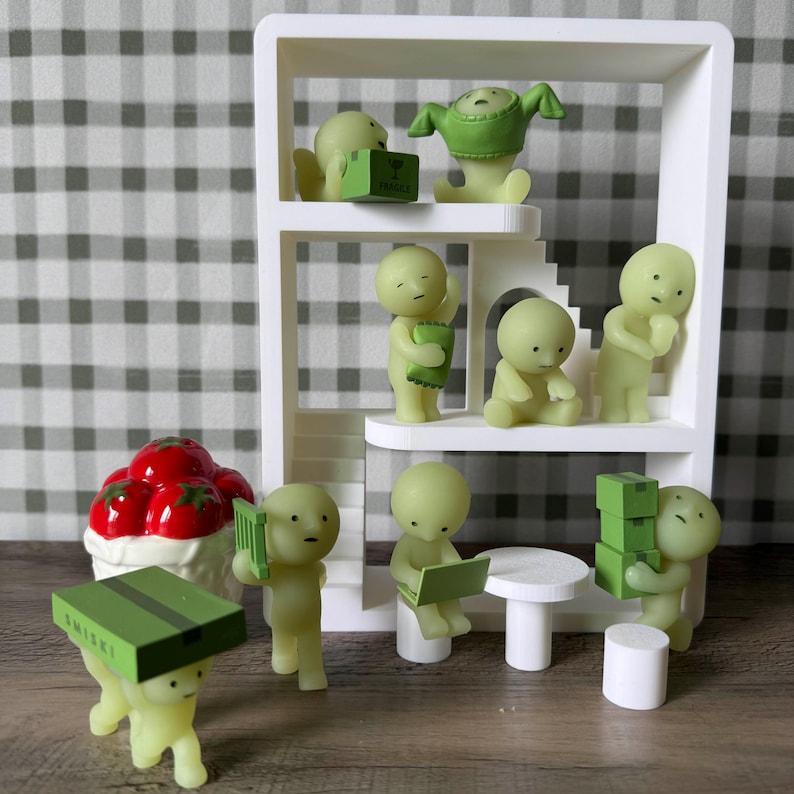 K&ouml;nnte beinhalten: Eine skurrile Anordnung kleiner, hellgr&uuml;ner Figuren auf einem wei&szlig;en, mehrst&ouml;ckigen Regal. Einige Figuren halten Kisten oder B&uuml;cher oder sitzen an einem kleinen Tisch. Ein dekoratives Dessert mit roten Fr&uuml;chten ist ebenfalls vorhanden.