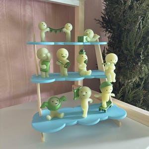May include: A three-tiered light blue cloud-shaped display stand holding nine small pale yellow figurines. The figures are depicted in various poses, including holding a green banner, sitting on a skateboard, and wearing a green pea-pod costume.