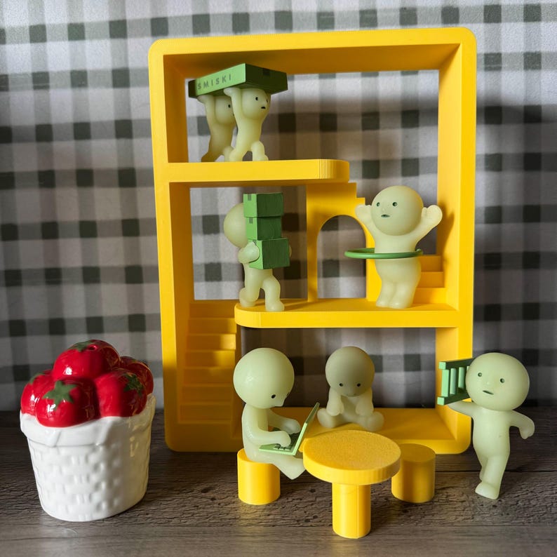 K&ouml;nnte beinhalten: Ein skurriles gelbes Miniaturgeb&auml;ude mit mehreren kleinen, leuchtenden Figuren, die verschiedenen Aktivit&auml;ten nachgehen. Einige Figuren tragen Kisten, andere scheinen zu arbeiten oder zu spielen. Ein wei&szlig;er Korb mit roten Tomaten steht daneben.