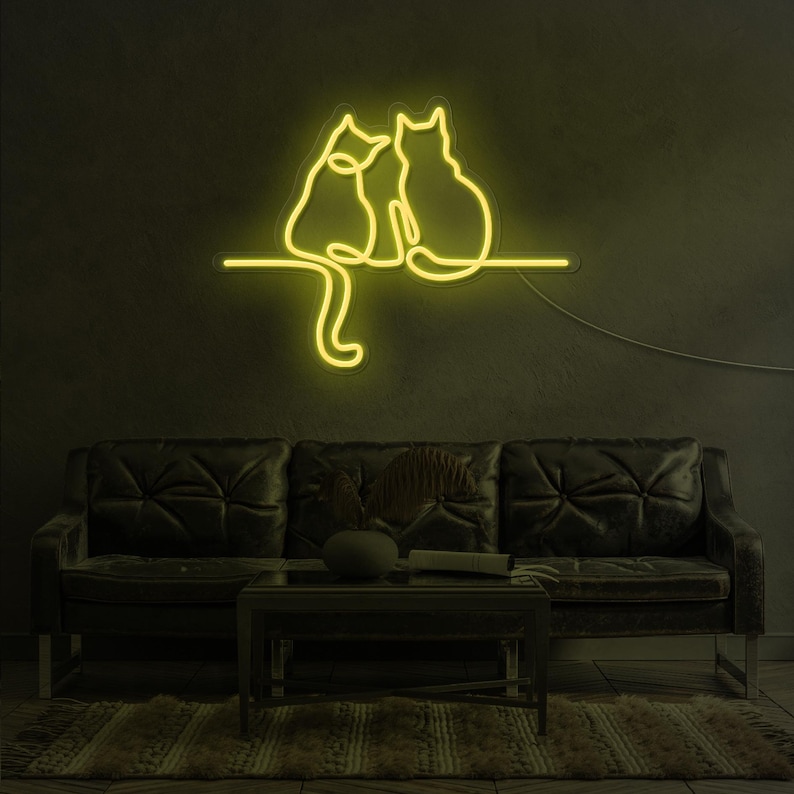 K&ouml;nnte beinhalten: Ein Neon-Schild, das zwei nebeneinander sitzende Katzen darstellt. Das Schild ist gelb und an einer dunklen Wand &uuml;ber einem schwarzen Ledersofa angebracht.