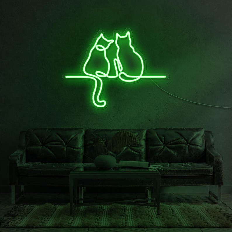 K&ouml;nnte beinhalten: Gr&uuml;nes Neon-Schild mit zwei Katzen, die nebeneinander sitzen. Das Schild ist an einer Wand &uuml;ber einem dunkelbraunen Ledersofa angebracht.