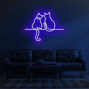 K&ouml;nnte beinhalten: Ein Neon-Schild, das zwei Katzen zeigt, die nebeneinander sitzen und ihre Schw&auml;nze ineinander verschlungen haben. Das Schild leuchtet in einem leuchtenden Lila und ist an einer Wand &uuml;ber einem dunklen Sofa angebracht.