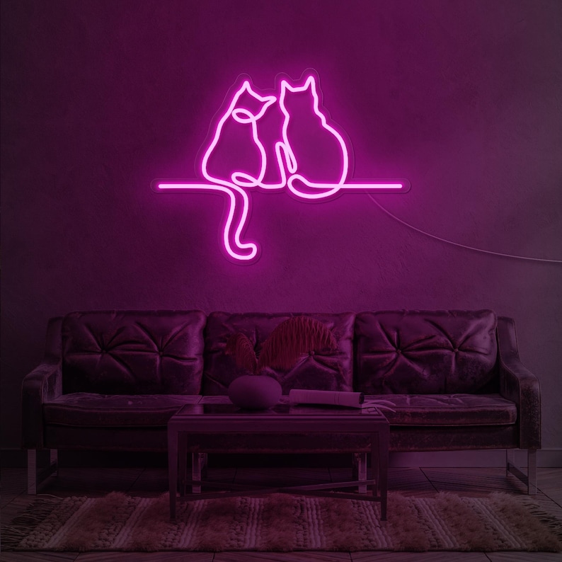K&ouml;nnte beinhalten: Pinkfarbenes Neon-Schild mit zwei nebeneinander sitzenden Katzen. Das Schild ist an einer Wand &uuml;ber einem dunkelbraunen Ledersofa angebracht.