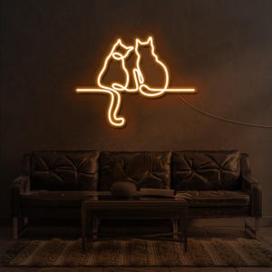 K&ouml;nnte beinhalten: Ein Neon-Schild, das drei Katzen zeigt, die auf einem Ast sitzen. Das Schild leuchtet in einem warmen Gelbton und ist an einer dunklen Wand montiert. Das Schild befindet sich &uuml;ber einem dunkelbraunen Ledersofa.