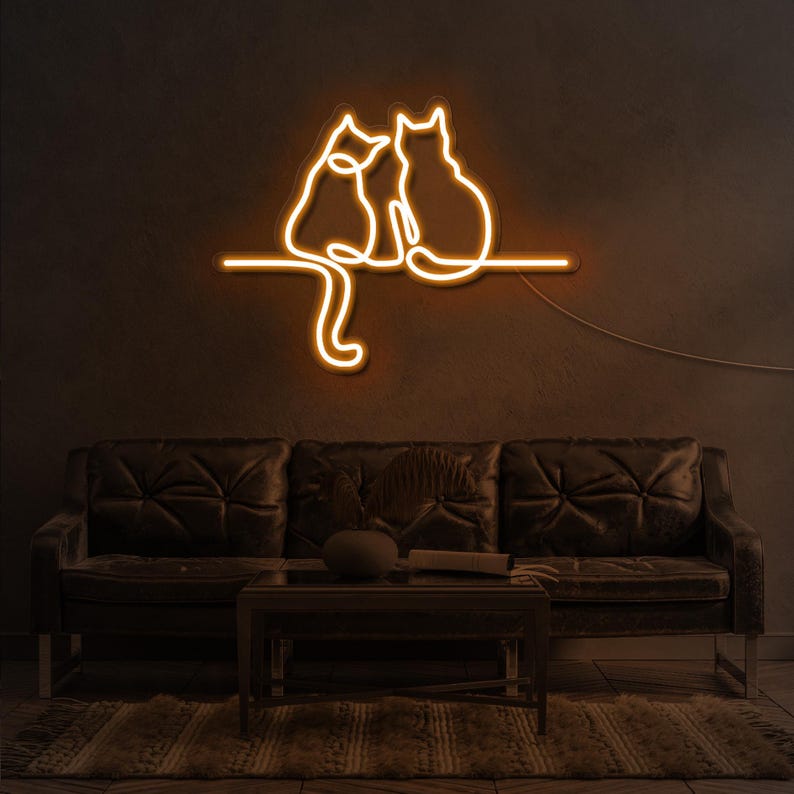 K&ouml;nnte beinhalten: Ein Neon-Schild, das drei Katzen zeigt, die auf einem Ast sitzen. Das Schild leuchtet in einem warmen Gelbton und ist an einer dunklen Wand montiert. Das Schild befindet sich &uuml;ber einem dunkelbraunen Ledersofa.