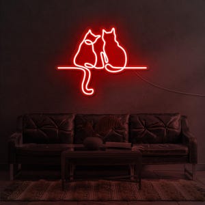K&ouml;nnte beinhalten: Rotes Neon-Schild mit zwei Katzen, die nebeneinander sitzen. Die Katzen sind in einer einzigen Linie umrissen und schauen in die gleiche Richtung. Das Schild ist an einer Wand &uuml;ber einem dunkelbraunen Ledersofa angebracht.