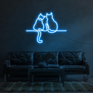 K&ouml;nnte beinhalten: Blaues Neon-Schild mit zwei Katzen, die nebeneinander auf einem Ast sitzen. Das Schild ist an einer dunklen Wand &uuml;ber einem schwarzen Ledersofa angebracht.