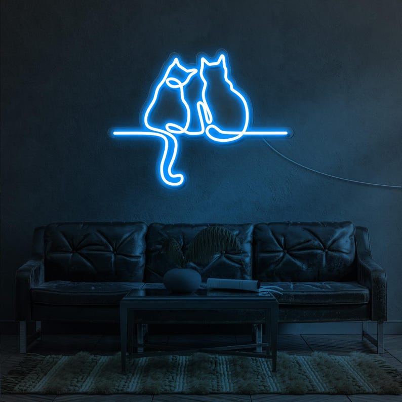 K&ouml;nnte beinhalten: Blaues Neon-Schild mit zwei Katzen, die nebeneinander auf einem Ast sitzen. Das Schild ist an einer dunklen Wand &uuml;ber einem schwarzen Ledersofa angebracht.