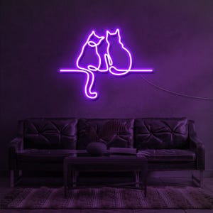 K&ouml;nnte beinhalten: Ein lilafarbenes Neon-Schild, das zwei nebeneinander sitzende Katzen darstellt. Das Schild ist an einer Wand &uuml;ber einem dunkelbraunen Ledersofa angebracht.