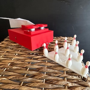 May include: A red mechanical device with two circular knobs and a white paper airplane attached, positioned next to a white triangular base holding ten small white bowling pins with red stripes. The base features the text "PAPER PLANE SYSTEMS".
