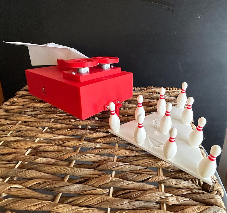 May include: A red mechanical device with two circular knobs and a white paper airplane attached, positioned next to a white triangular base holding ten small white bowling pins with red stripes. The base features the text "PAPER PLANE SYSTEMS".