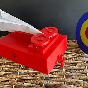 May include: A bright red paper airplane launcher featuring two circular wheels on top and a small trigger mechanism on the side. A white paper airplane is positioned in the launcher, and a circular target with blue, yellow, and red rings stands nearby.